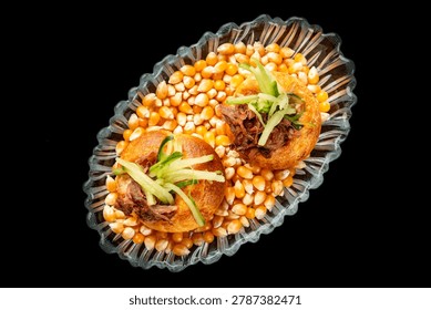 Two puff pastries filled with pulled beef and julienned green onions, served on a bed of dry corn kernels in a glass bowl against a dark background.