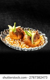 Two puff pastries filled with pulled beef and julienned green onions, served on a bed of dry corn kernels in a glass bowl against a dark background.