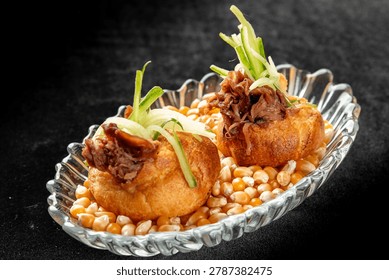 Two puff pastries filled with pulled beef and julienned green onions, served on a bed of dry corn kernels in a glass bowl against a dark background.
