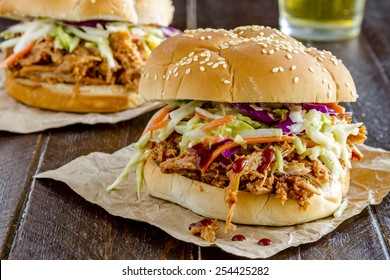 Two pulled pork barbeque sandwiches with coleslaw sitting on wooden table with glass of beer