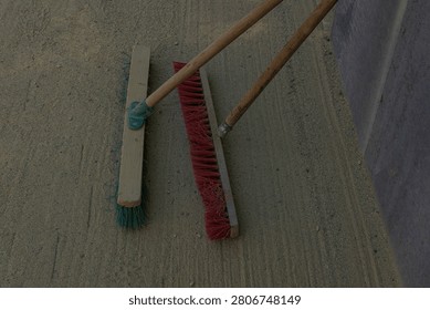 Two push brooms resting on sandy ground near a concrete structure at a construction site. Cleaning tools used for sweeping sand, dust, and debris during construction and maintenance work