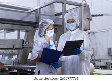 Dos inspectores de control de calidad vistiendo ropa protectora, mascarillas y guantes revisan las notas sobre los Portapapeles mientras monitorean la producción de alimentos en una fábrica.