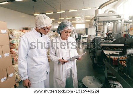 Two quality professionals in white sterile uniforms checking quality of salt sticks while standing in food factory.