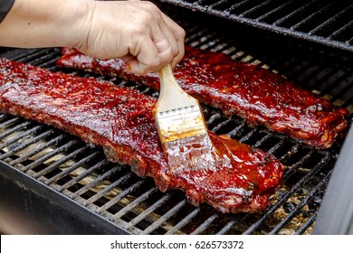 Two Racks Of Pork Bbq Ribs Sitting On Grill With Hand Applying Barbeque Sauce With Brush