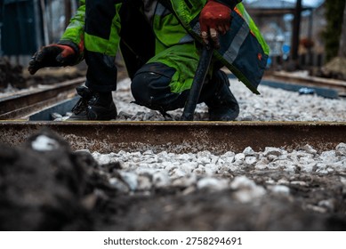 Two rail workers crouch and adjust steel with a crowbar on fresh ballast. Gloved hands, worn boots, tools, and sleepers are visible in a cold daytime rail corridor.
