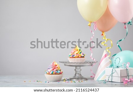 Two Rainbow Birthday cupcakes with presents, hats and colorful balloons over light grey background. Scene from a birthday party!