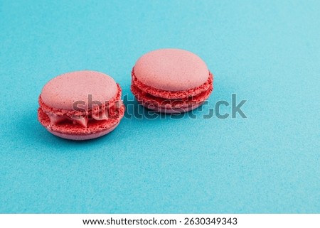 Two raspberry macarons sit invitingly on a bright turquoise surface, showcasing their smooth texture and vibrant color, tempting any dessert lover nearby.