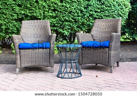 two ratan chairs  with blue cushions outside with a small decorative table on brick with green bush background
