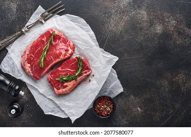 Two raw steak New York with rosemary and spices on a piece of parchment paper on old dark stone background. Top view. Mock up.