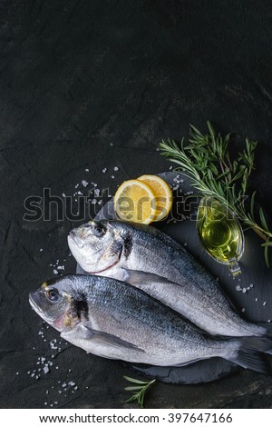 Two ready to cook raw bream fish with herbs, lemon and olive oil on stone slate board over black textured background. With copy space. Top view