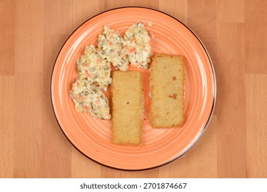 Two rectangular breaded fish filets served with a portion of classic French salad (Croatian way) on an orange-rimmed plate on a light wood surface.