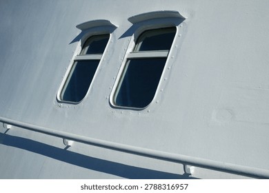 Two rectangular marine windows with protective metal eyebrows installed on a clean white steel ship hull under bright direct sunlight.