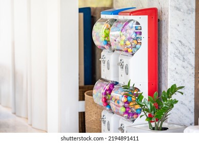 Two red and blue coin operated gachapon cabinets which inside is filled with colorful capsules are located on a light brown marble corridor inside the building.
