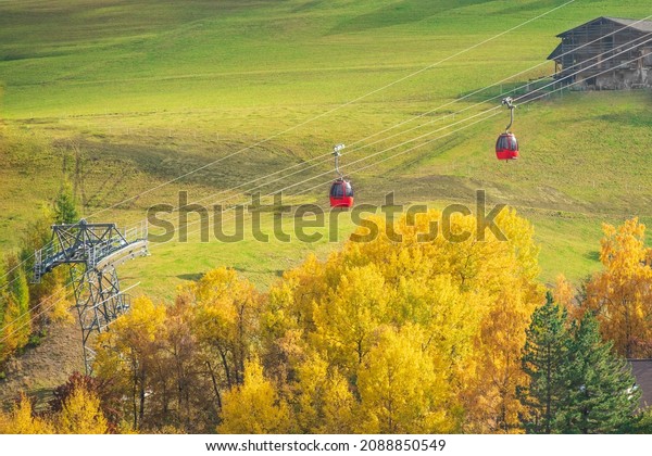 Two red cable car cabins hang over a green field\
and yellowed trees