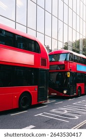 Two red double-decker buses parked on a london street, reflecting in the glass facade of a modern office building