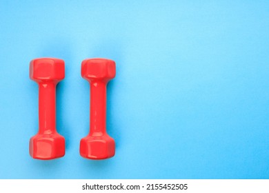 Two Red Dumbbells On A Blue Background In The Corner Of The Frame. Color Contrast Blue And Red. Sports Equipment On A Blue Background