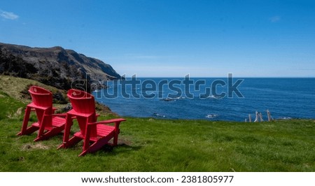 Two red empty chairs on lush green grass on the seashore in Newfoundland, Canada