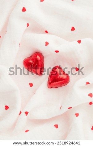 Two red heart shaped chocolate candies on white fabric background with tiny hearts print