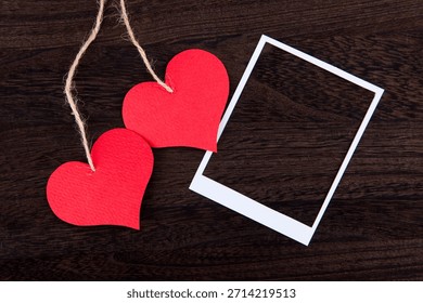 Two red hearts with string hang beside an empty white frame on a wood surface