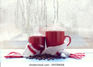 Two Red Mugs In A Scarf Stand On A Table In The Background Of A Window With Dripping Rain Drops / Cozy Aroma Of Coffee