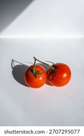 Two red tomatoes connected by a green vine, shining in the sun on a white background.