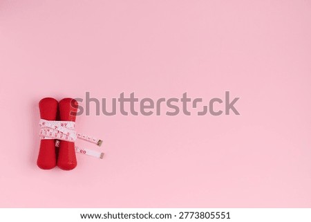 Two red weights are tied with a measuring tape on a pink surface showing a focus on fitness and health.