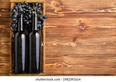 Two Red Wine Bottles Composition On Brown Wooden Table. Red Wine Bottles In Box On Black Ripe Grapes On Wooden Table. Old Collection Wine Label Template Top View With Copy Space