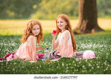 Two red-haired curly happy girls children play picnic on grass among white clover flowers