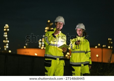 Two refinery engineers communicate via walkie-talkie while reviewing data on a tablet at a plant site at night. Safety protocols, and efficient nighttime operational management.