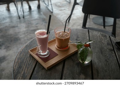 Two refreshing drinks, an iced coffee and a pink smoothie, served on a wooden tray on a rustic cafe table in a modern indoor setting.