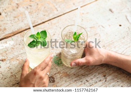 Two refreshing drinks in transparent relief glasses with ice, white straws and mint leaves are in the hands of a man and woman with a wedding ring on her finger in the background of old wooden table