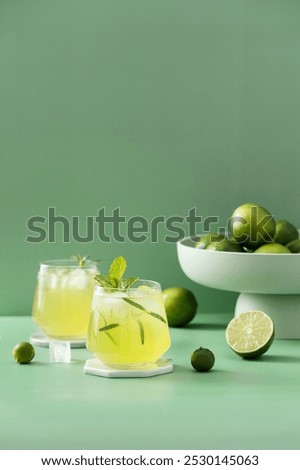 Two refreshing lemon drinks with mint sprigs on a clear surface; a bowl filled with whole limes sits in the background alongside sliced lime.