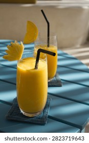 Two refreshing tropical fruit smoothies served in tall glasses with black straws, garnished with fresh pineapple slice and orange wheel, placed on a blue wooden Slatted table under natural sunlight.