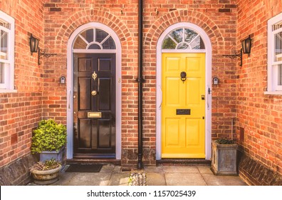 Two residential front doors, one yellow, one black with a drain pipe down the middle. The walls are red brick there are two side windows and lunette arches over the doors