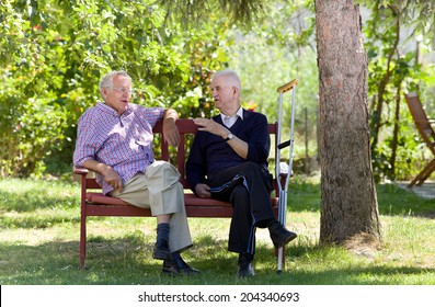 Two Retired Men Sitting On Bench In Park And Talking