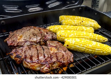 Two ribeye steaks and corn on the cob cooking over flames on grill