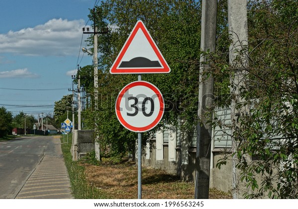 two road signs speed limit and\
speed bump on an iron pole against a green tree and blue\
sky