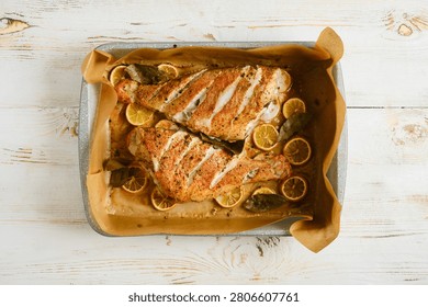 Two roasted red snapper are placed on baking tray, surrounded by lemon slices and herbs. Dish is golden brown and prepared in oven