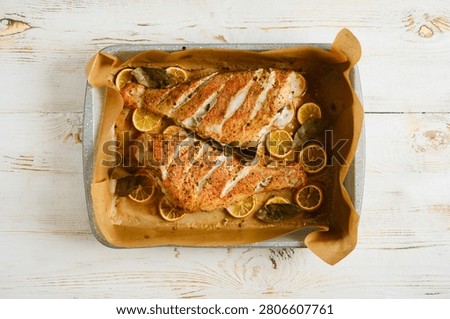 Two roasted red snapper are placed on baking tray, surrounded by lemon slices and herbs. Dish is golden brown and prepared in oven