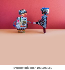 Two robots greet each other. Handshake of cybernetic mechanical cyborgs. Creative design robotic toys, red wall brown floor background. Copy space.
