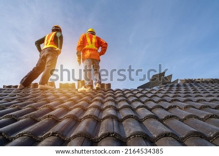 Two Roofer workers in protective uniform wear and gloves, using air or pneumatic nail gun and installing Concrete Roof Tiles on top of the new roof, Concept of residential building under construction.
