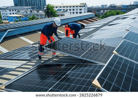 Two rooftop solar energy workers in protective safety gear skilfully carry a panel in tandem over the roof crest in silhouette high on an industrial warehouse roof in a Thai city.