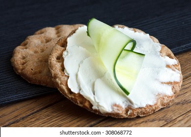 Two round rye crispbreads on wood with black napkin. Top one spread with cream cheese and cucumber slice.