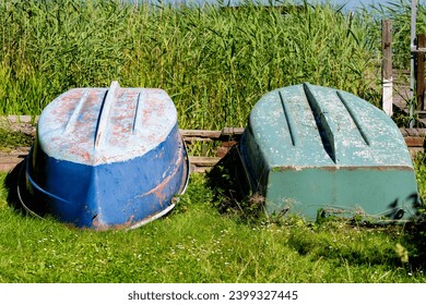 Two rowing boats lying upside down on grass on the shore. The paint on the boats is already very chipped. You can see reeds in the background and the lake further back