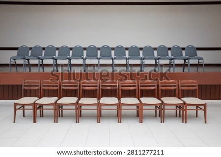 Two rows of neat, elegant chairs in a spacious, pristine, yet empty room, arranged in two rows. One row is positioned on top of a raised pulpit platform, while the other is situated directly below at its foot. One row is a sleek slate gray, and the other is a warm coffee-and-cream brown.