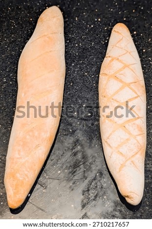 Two rustic loaves of artisan bread displayed side by side on a dark textured surface, showcasing distinct scoring styles—one with diagonal slashes and the other with a crisscross pattern.