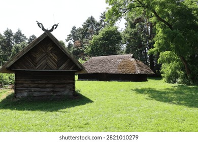 Two rustic log cabins with distinctive thatched roofs stand in a vibrant green field surrounded by lush trees under a bright sky