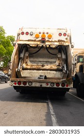 Two rusty garbage trucks are parked on an urban street, showcasing their rear ends with open compartments.
