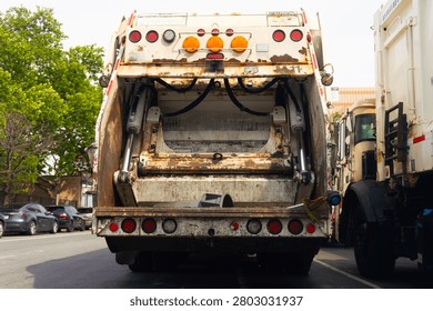 Two rusty garbage trucks are parked on an urban street, showcasing their rear ends with open compartments.

