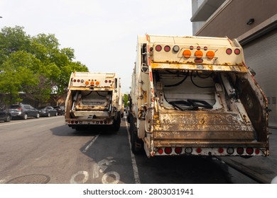 Two rusty garbage trucks are parked on an urban street, showcasing their rear ends with open compartments.

